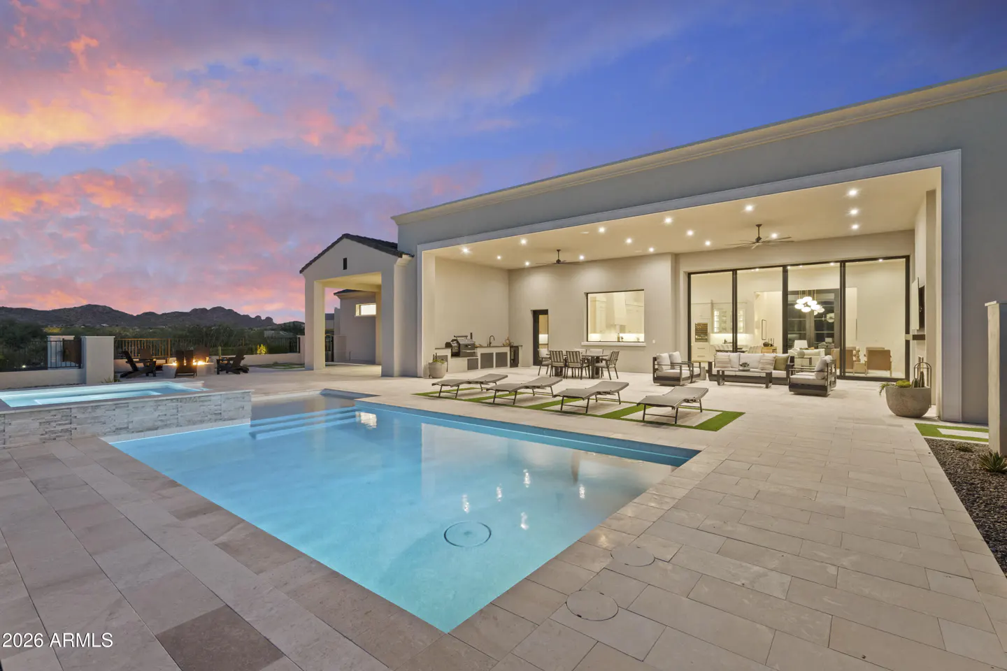 Luxury backyard with a glowing pool, stone patio, covered outdoor living area, and sunset mountain views.
