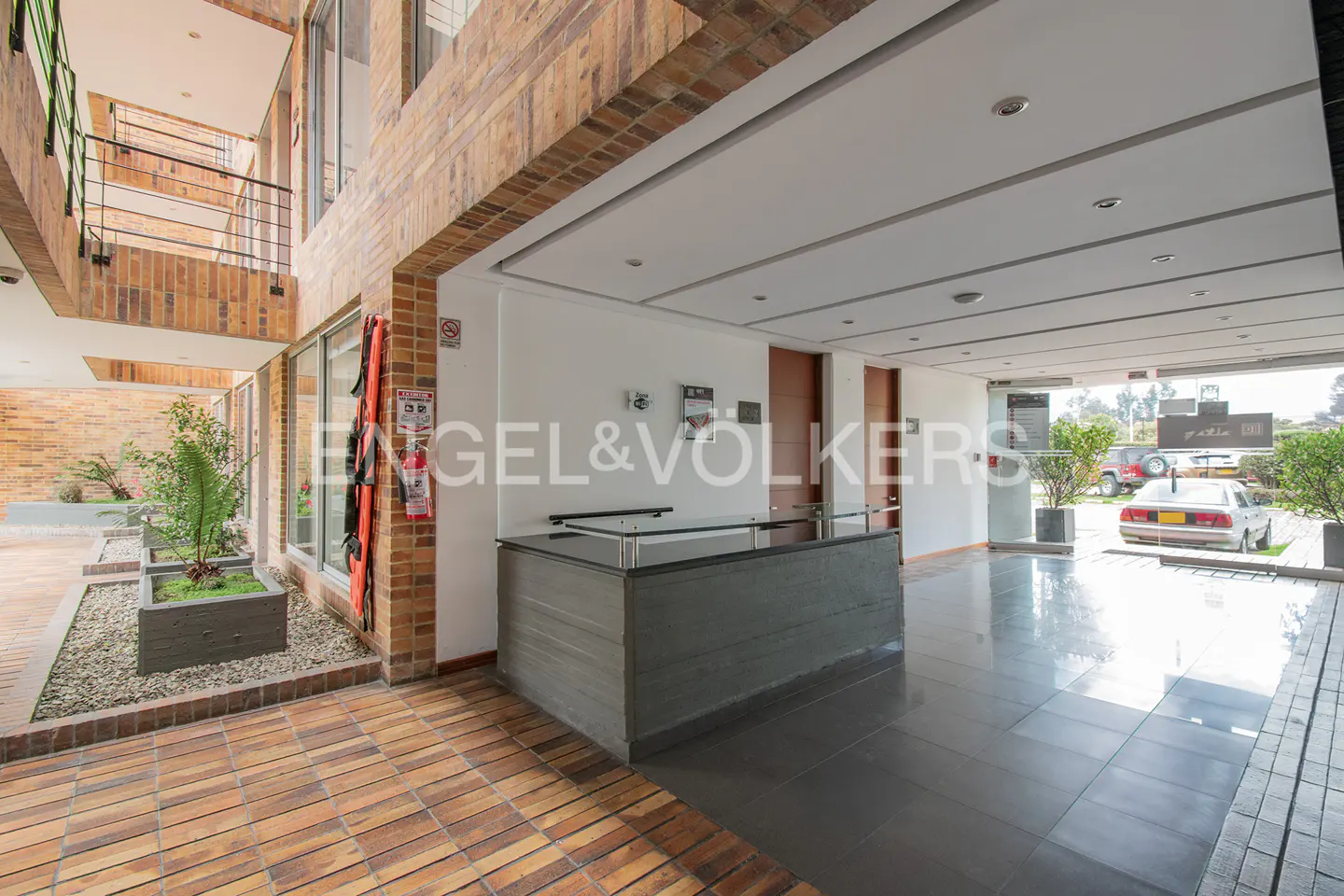Lobby of a building with a gray reception desk, brick walls, and tile floors. The Engel & Volkers logo is visible. Cars are parked outside.