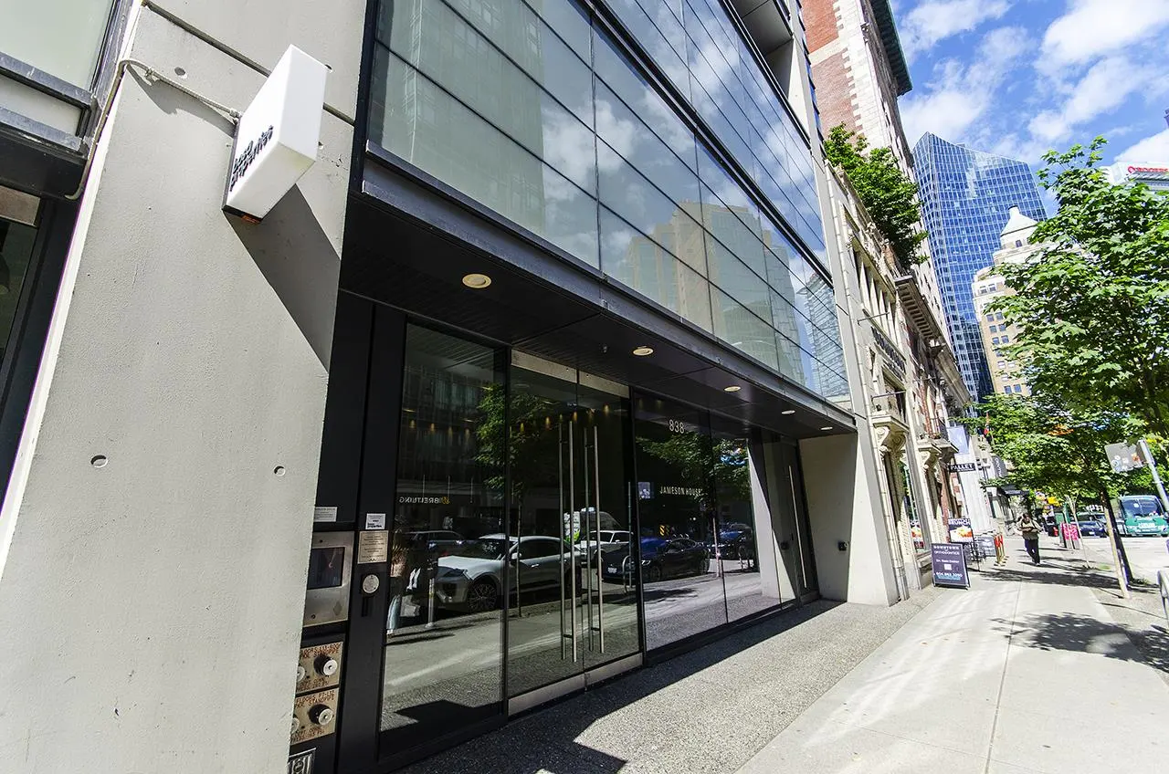 Exterior of a modern building with glass windows and a gray concrete facade. A white sign is mounted on the building. The sidewalk is visible in the foreground.