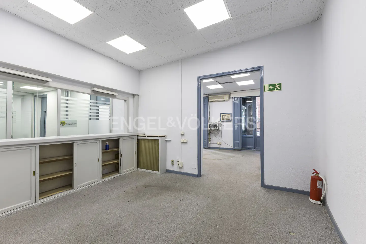 Empty office space with white walls, gray carpet, and fluorescent lighting. A doorway leads to another room. A red fire extinguisher is visible.