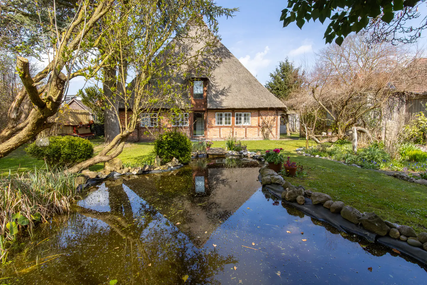Exterior view of a thatched-roof house with a pond in the front yard on a sunny day. The house is brick with white-framed windows.