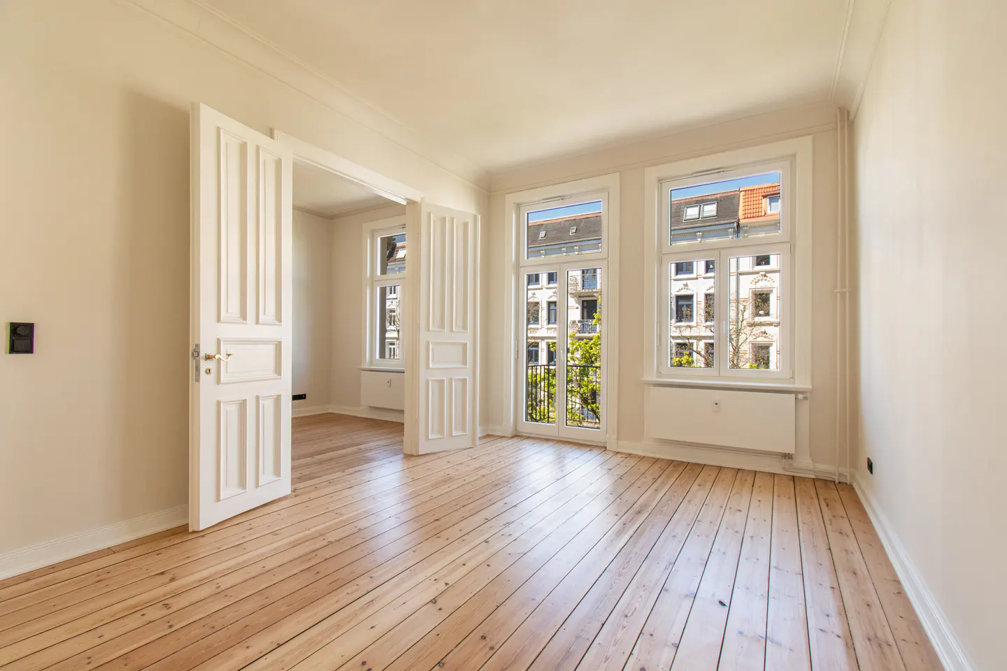 Bright, empty room with light wood floors, white walls, and two large windows showing a city view. A white door is open to another room.