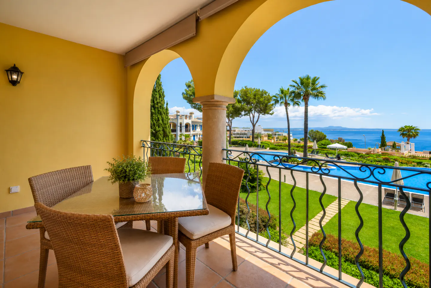 Covered balcony with wicker dining set overlooking a swimming pool, lush gardens, and the blue Mediterranean sea.