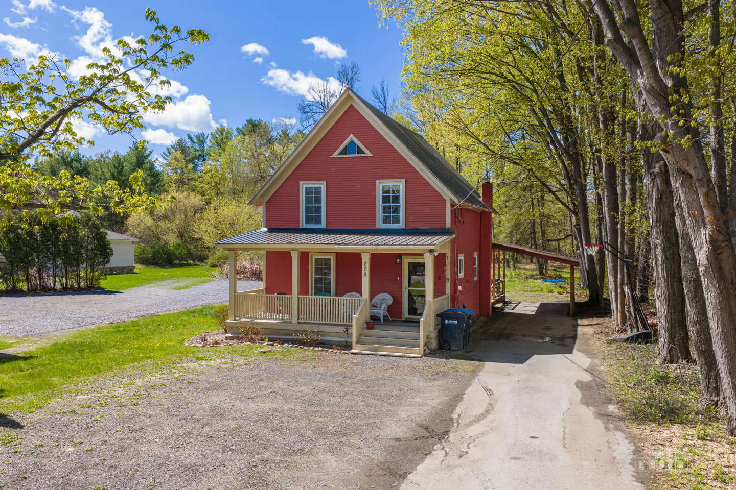 A two-story red house with a porch and a gravel driveway surrounded by trees.