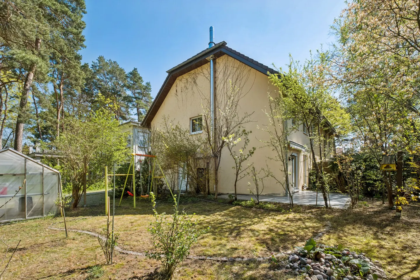 A tan two-story house with a brown roof, a metal chimney, and a green lawn with trees and a greenhouse.