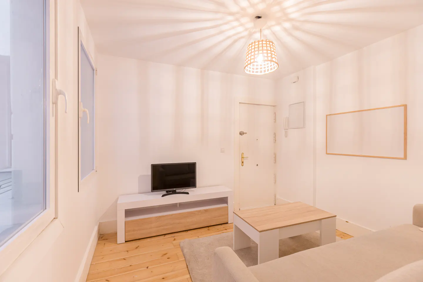 Bright living room with white walls, wood floors, and a beige sofa. A TV sits on a white and wood stand. A woven pendant light hangs from the ceiling.
