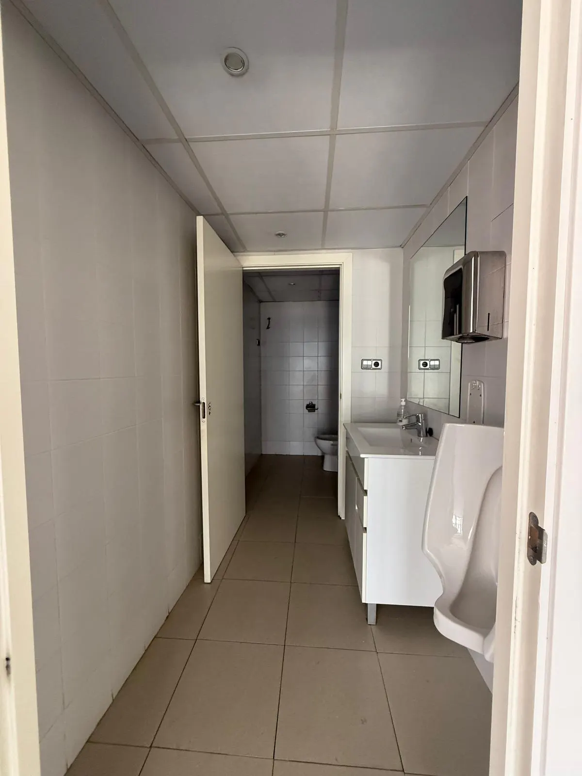 A bright, empty commercial bathroom with white walls, a urinal, and a sink. A doorway leads to a tiled toilet area.