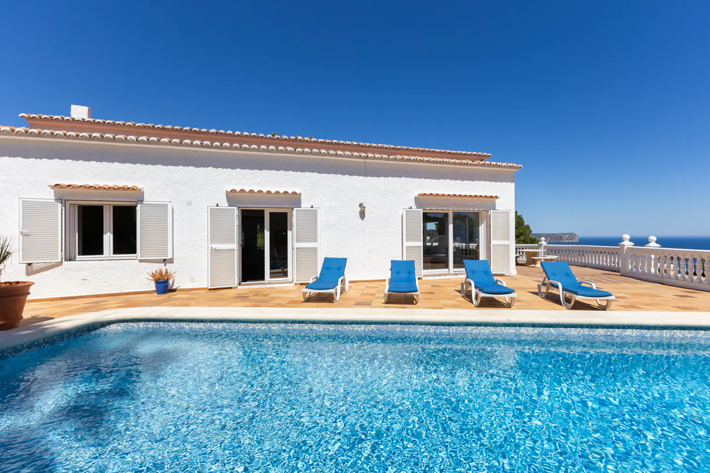 A white villa with a pool and blue lounge chairs under a clear blue sky.