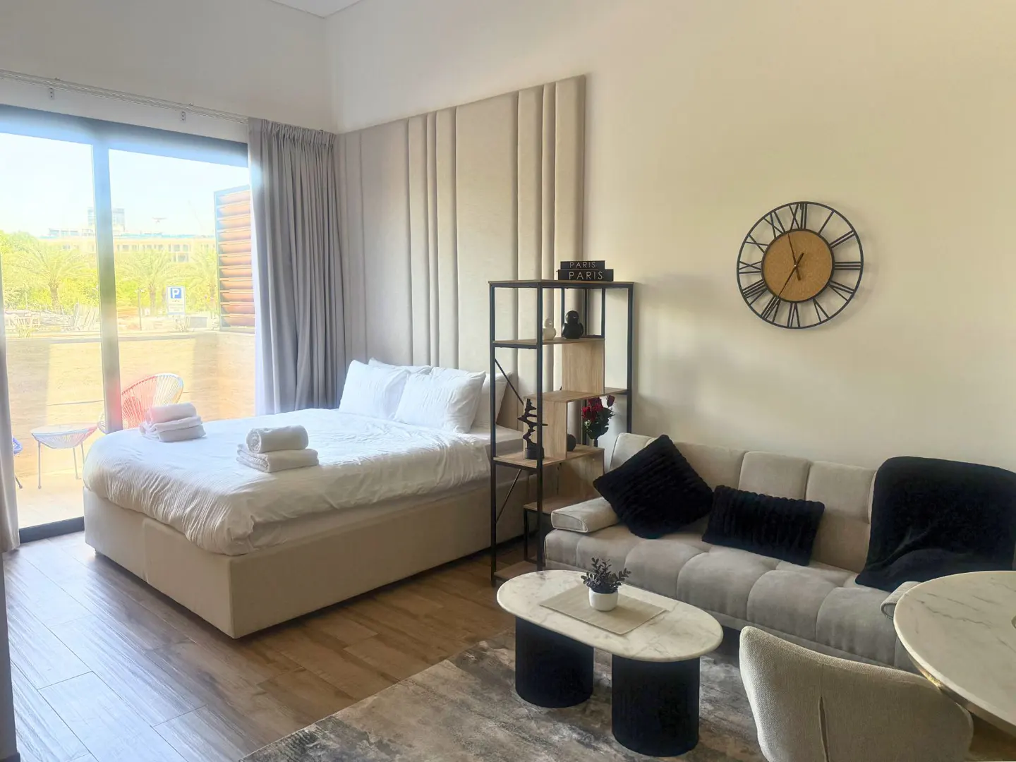 Studio apartment interior with a bed, sofa, coffee table, and balcony with outdoor seating. A clock hangs on the wall.