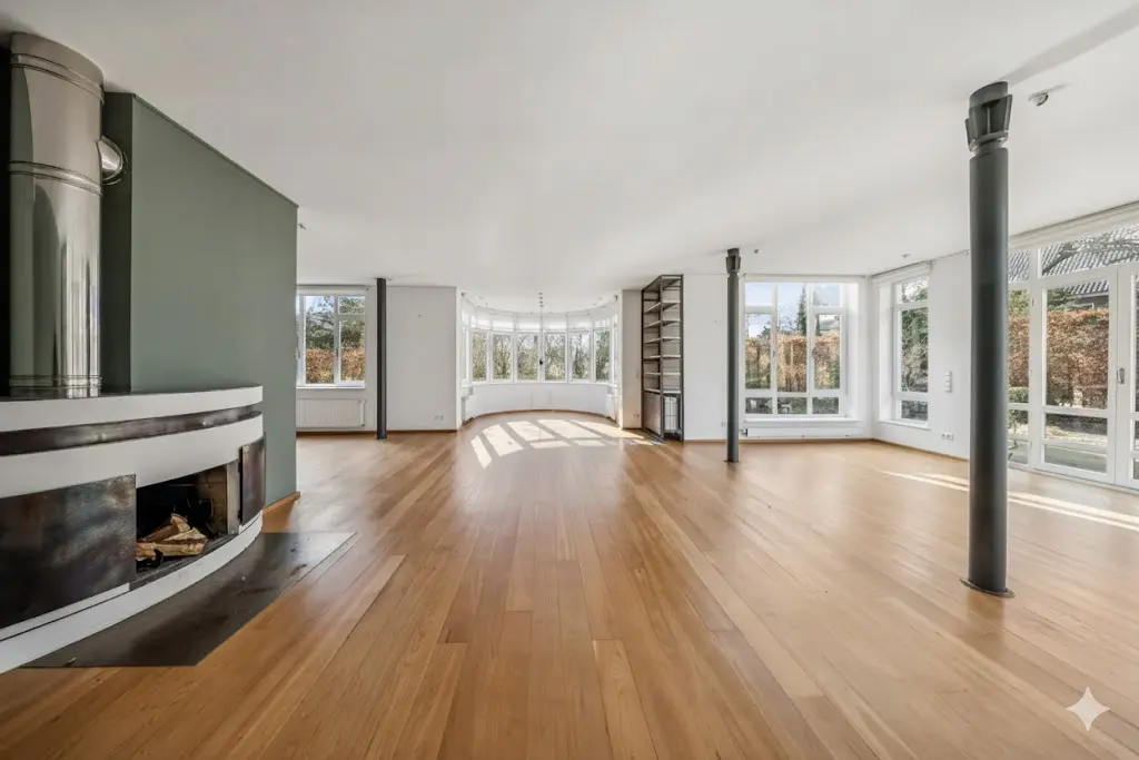 An empty living room with hardwood floors, a fireplace, large windows, and gray support columns.