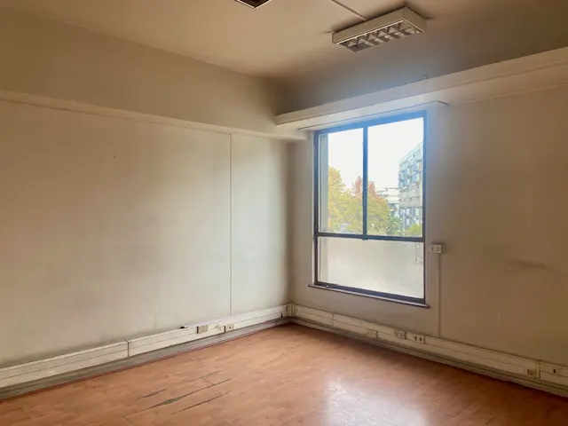 Empty office space with wood floors, beige walls, and a large window showing a city view.