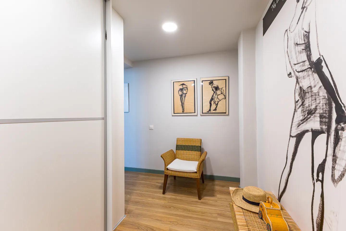 Hallway with a white closet, wood floor, and gray walls. A wicker chair sits beneath framed art. A bench holds a straw hat and yellow toy car.