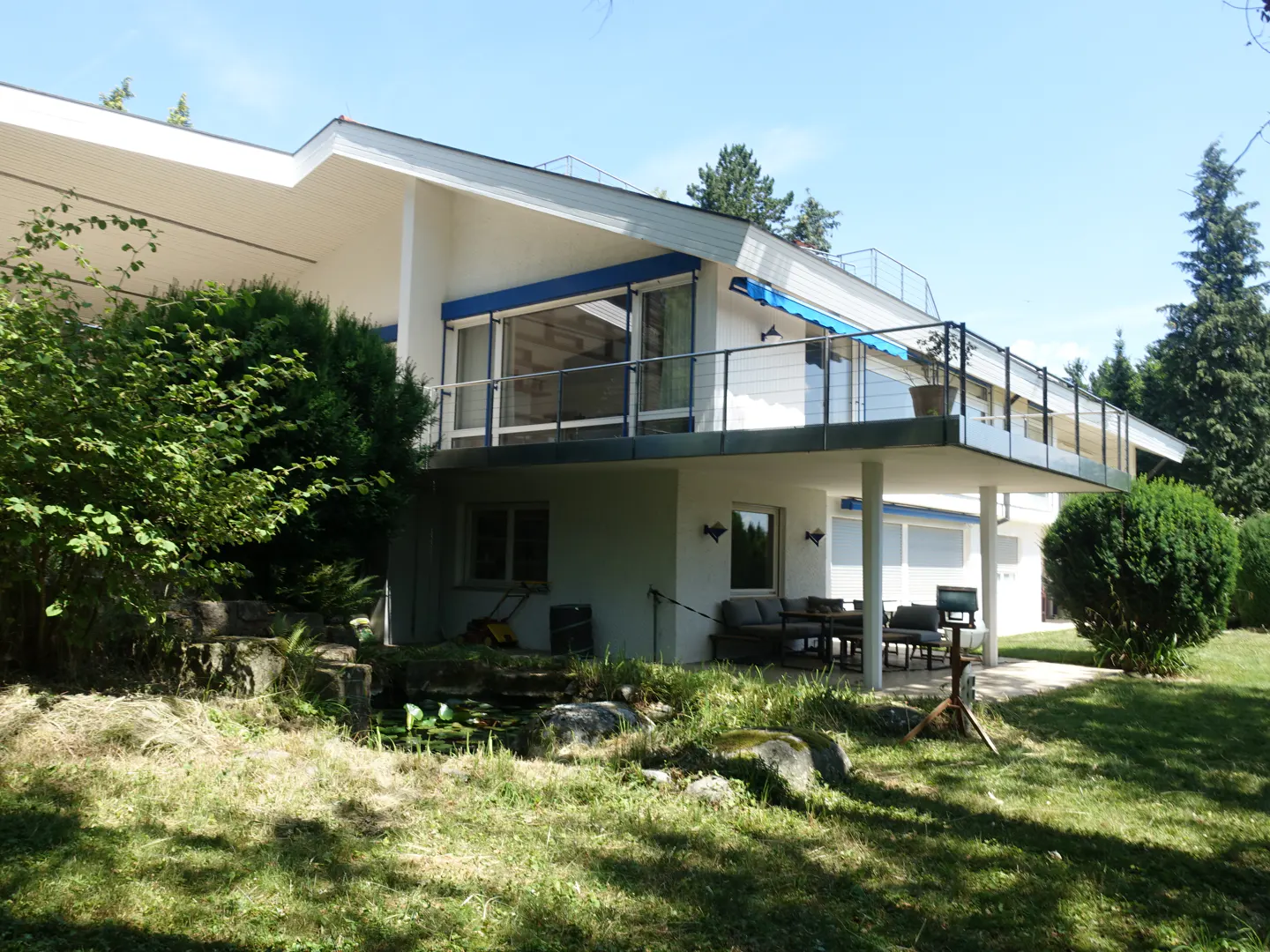 Modern white two-story house with a large balcony, blue awnings, a garden pond, and a patio area on a sunny day.