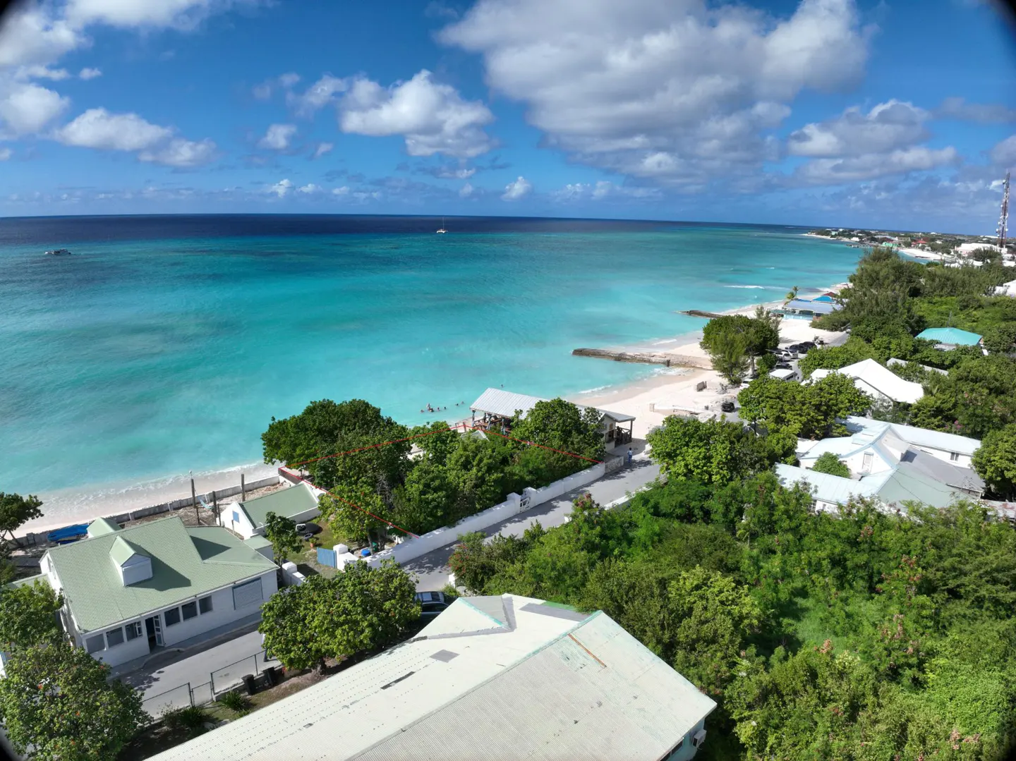 Coastal view of turquoise ocean meeting a white sand beach, lined with green trees and white-roofed buildings under a blue sky with scattered clouds.