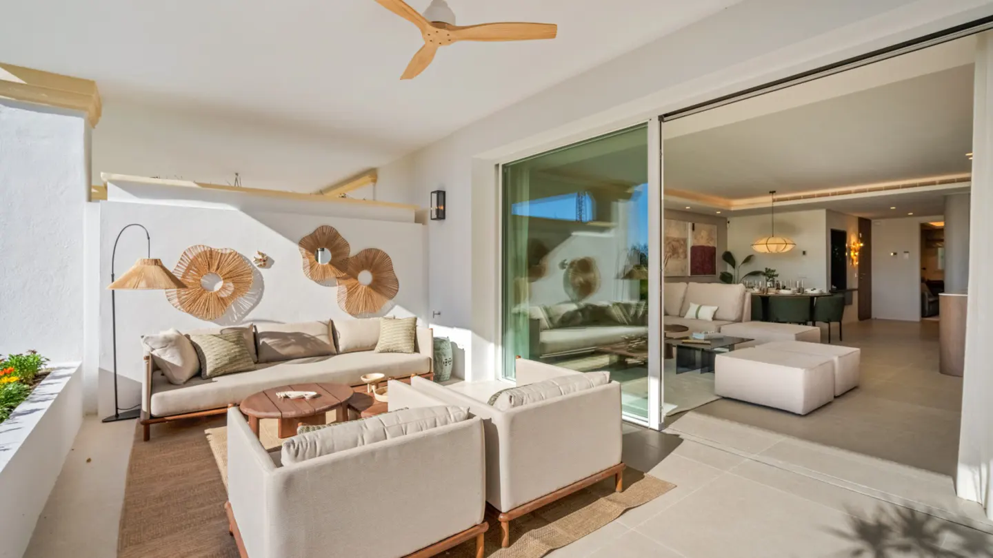 A bright, modern patio with a beige sofa, two white armchairs, and a wooden coffee table. Wicker art adorns the white wall. Sliding glass doors open to a living room.