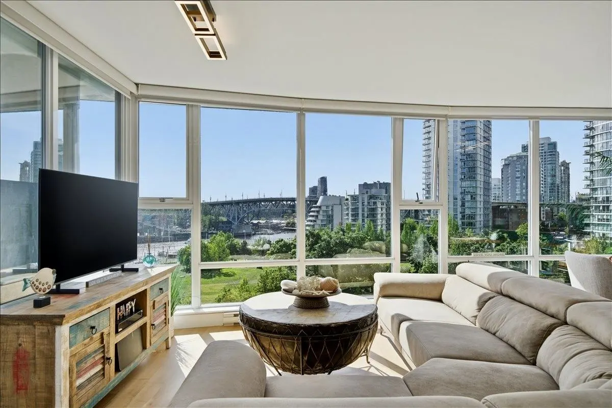 Bright living room with a beige sectional sofa, a round coffee table, and a TV on a rustic cabinet. Large windows offer city and bridge views.