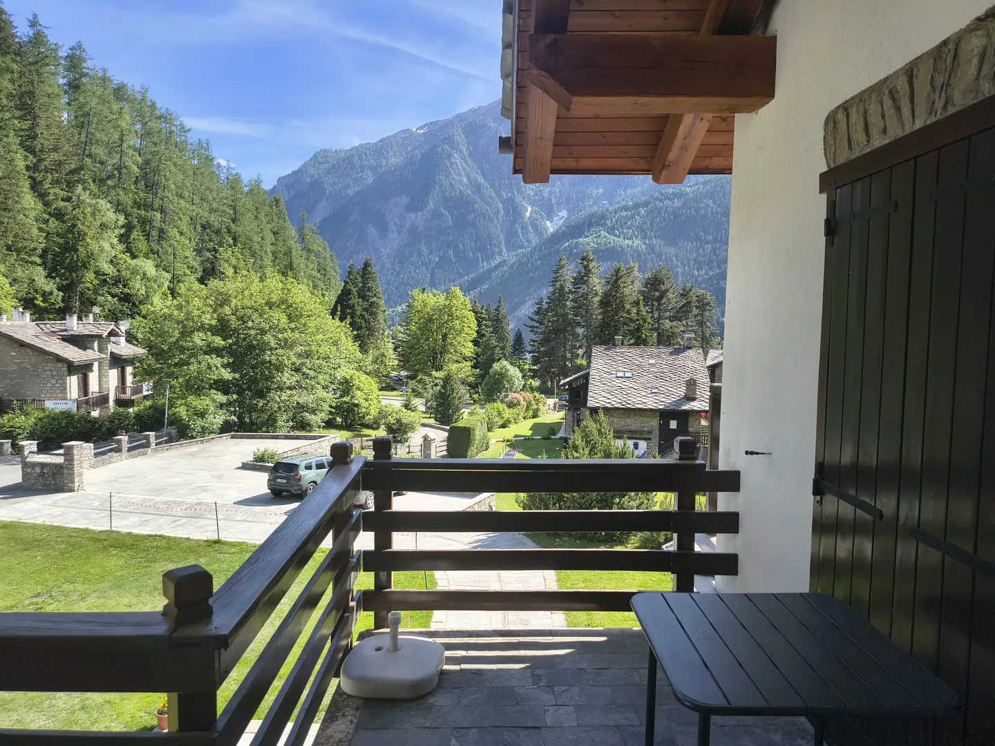 View from a balcony overlooking a lush green valley, stone houses, and towering mountains under a clear blue sky.