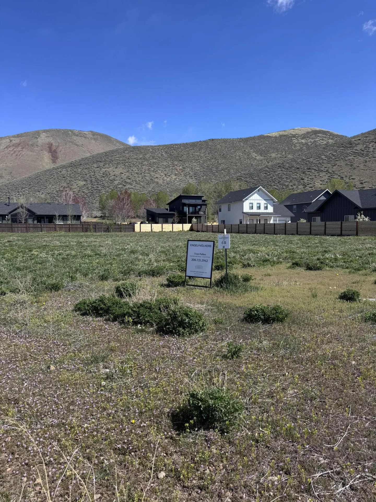 Vacant lot with a real estate sign in front of houses and a hill under a blue sky.