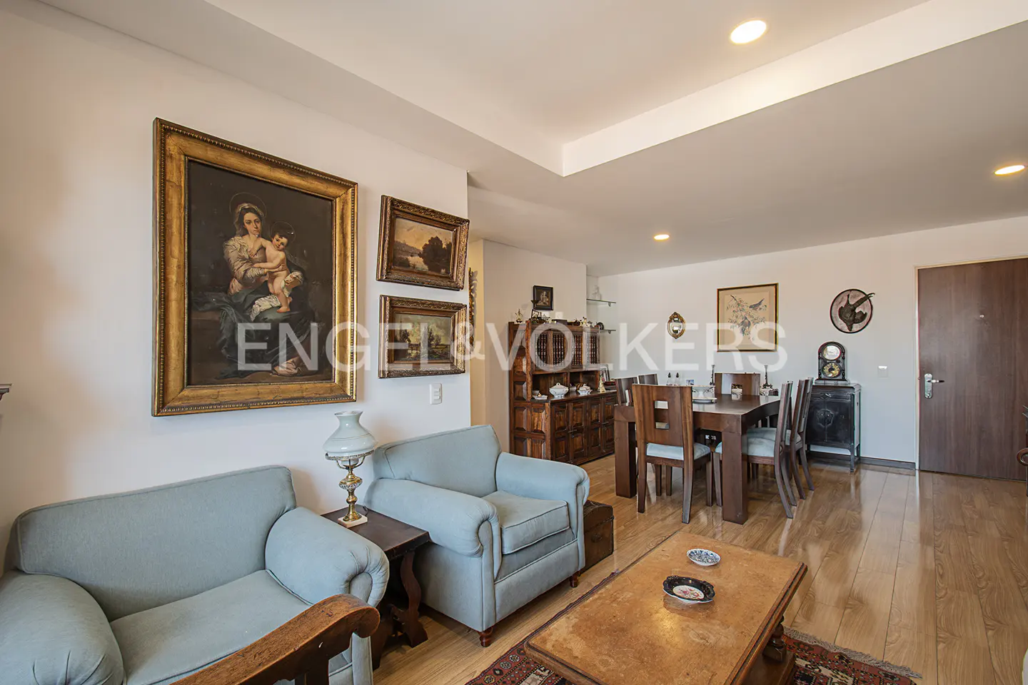 Living room with light blue armchairs, wooden floors, and framed paintings on the wall. A dining table is visible in the background.