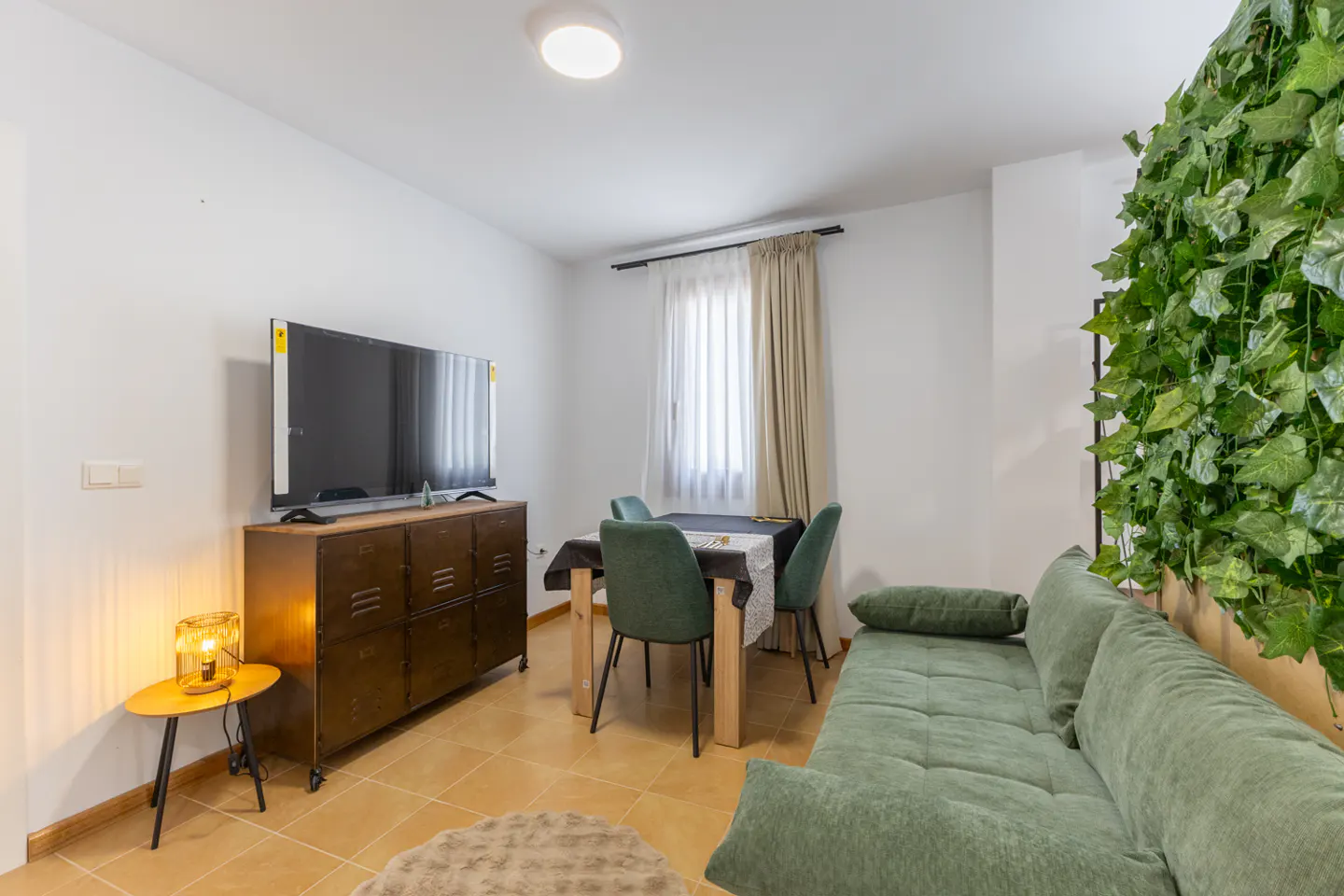 Living room with green sofa, dining table with four chairs, TV on a brown cabinet, and a wall covered in green ivy.