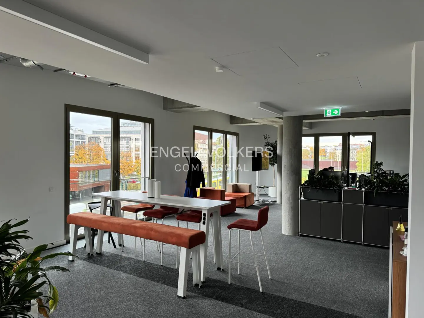 Bright office space with a long white table, orange bench and chairs, and large windows overlooking a cityscape.