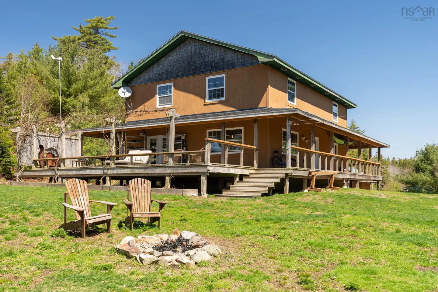 Two-story brown house with a large deck, two wooden chairs, and a fire pit in the yard.