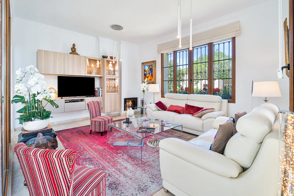 Bright living room with white sofas, red rug, and a glass coffee table. A TV is mounted above a cabinet, and a window lets in natural light.