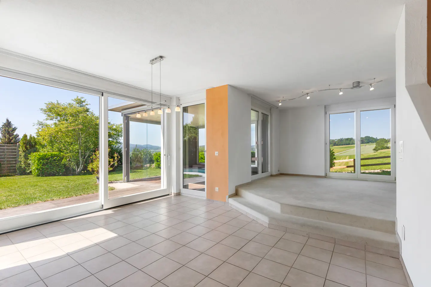 Bright, open living area with tiled floors, a raised carpeted platform, and large glass doors overlooking a green garden.