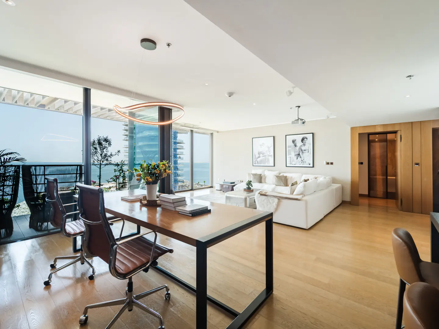 Bright, modern apartment interior with a wood desk, leather chairs, and a white sofa. Ocean view through large windows.