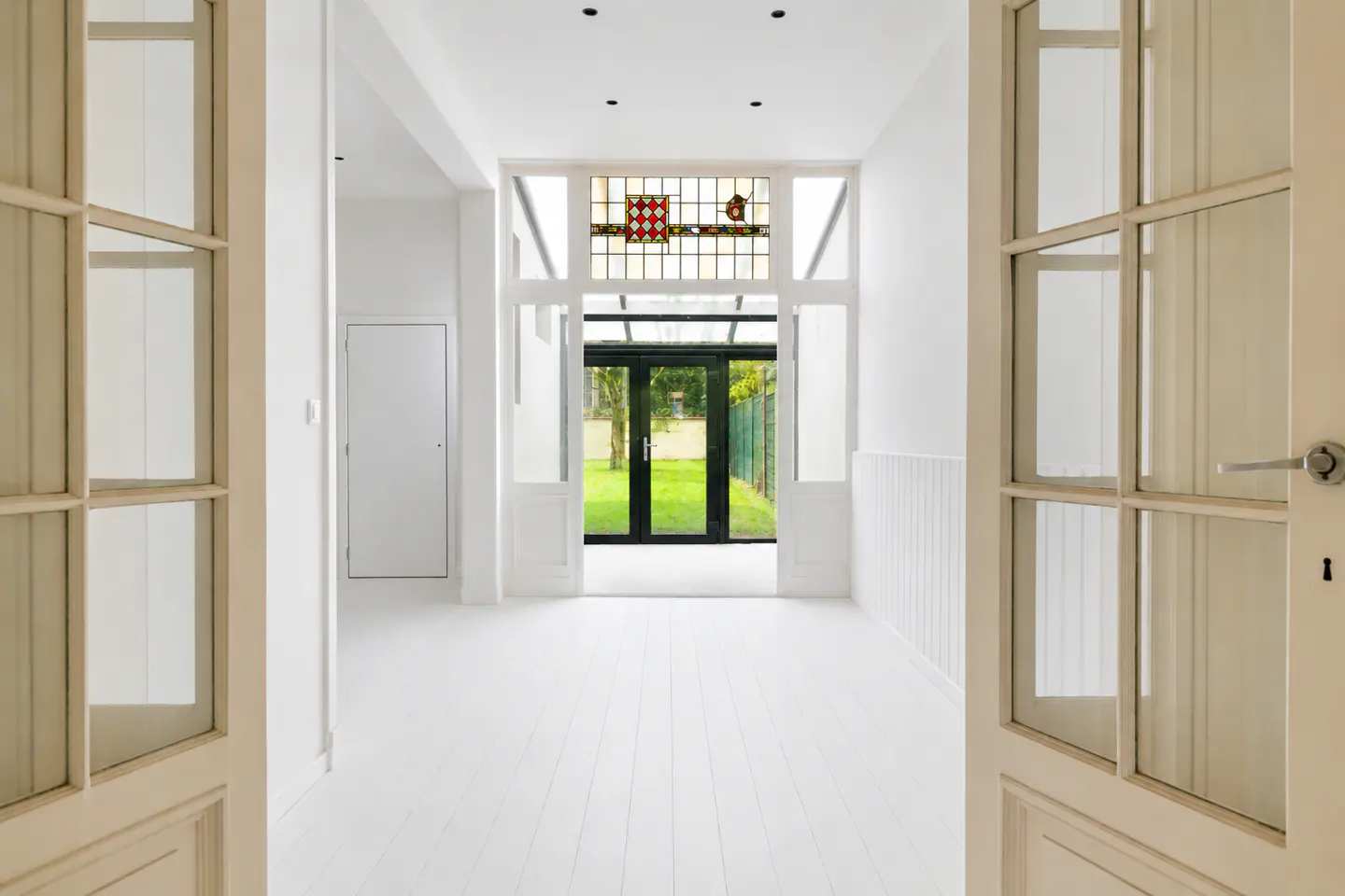 Bright white hallway with open french doors. Stained glass window above black framed doors leading to a green lawn.