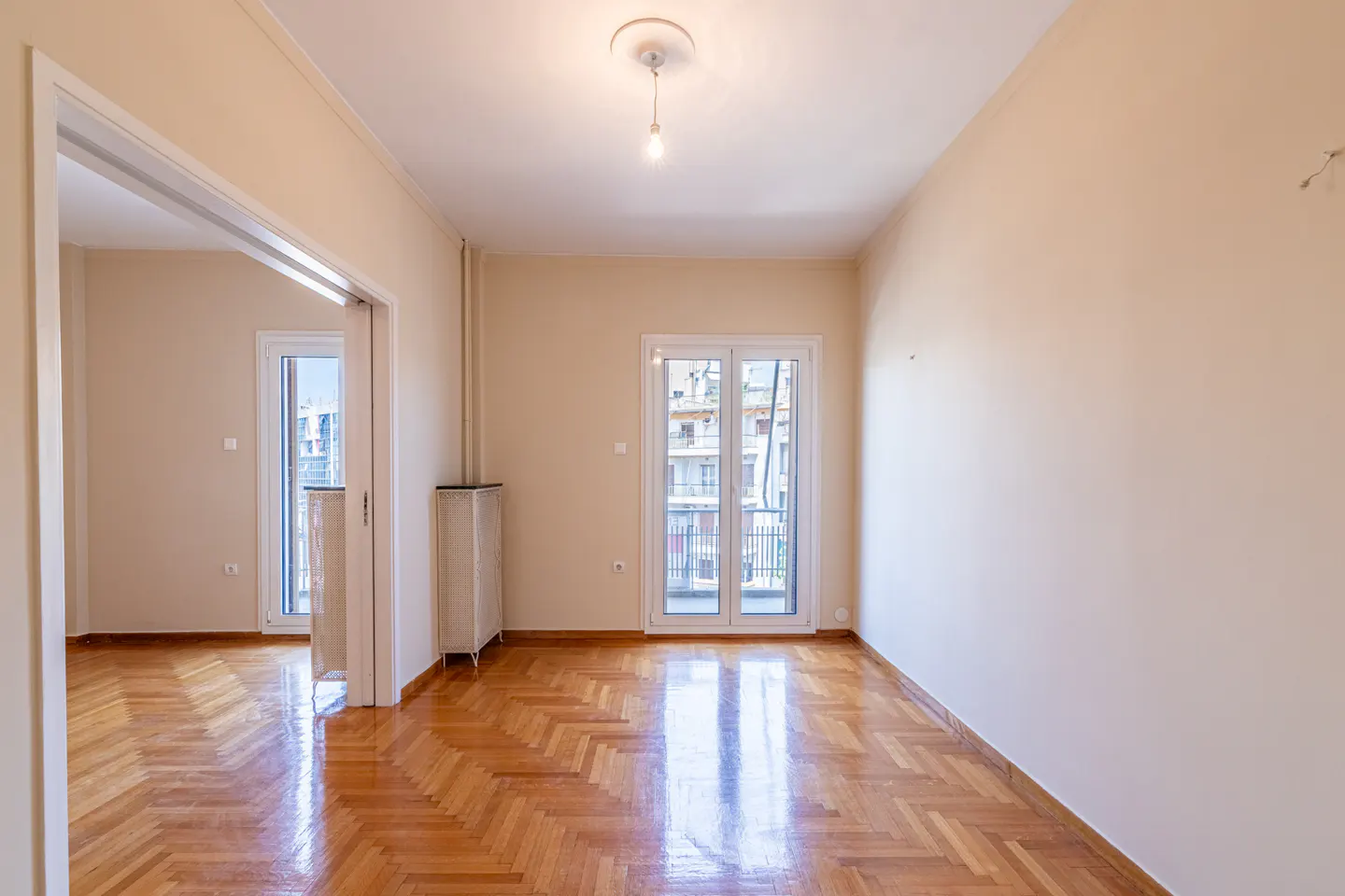 Empty room with beige walls, polished herringbone wood floors, and glass doors leading to a balcony.