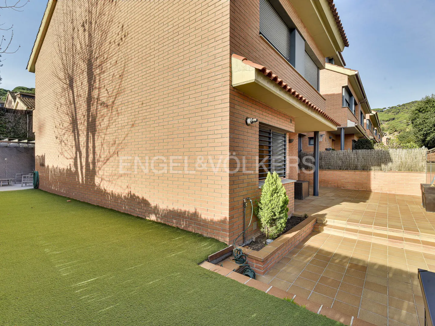 Exterior view of a brick townhouse with a green lawn and tiled patio. A tree shadow is visible on the brick wall.