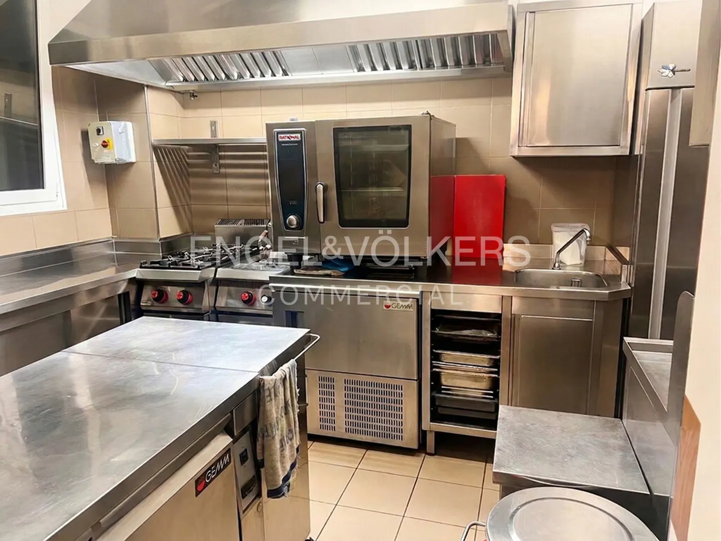Commercial kitchen with stainless steel appliances, including a stove, oven, and sink. The walls are tiled in a neutral color.