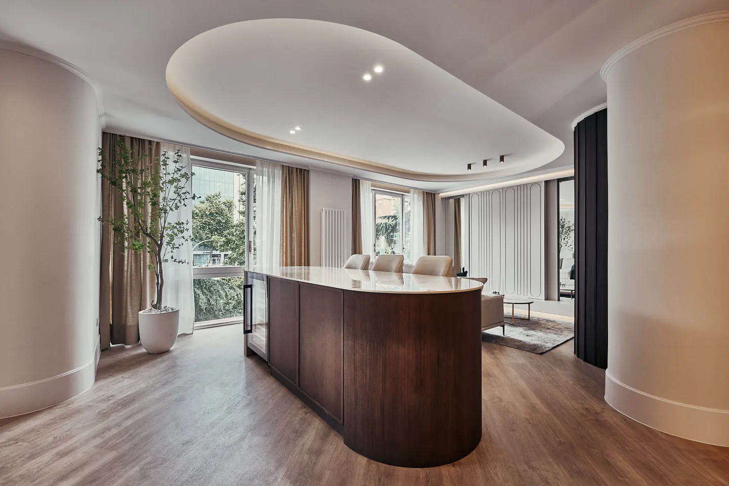 A modern living room with a wood floor, a brown island with a white countertop, and large windows with sheer curtains.