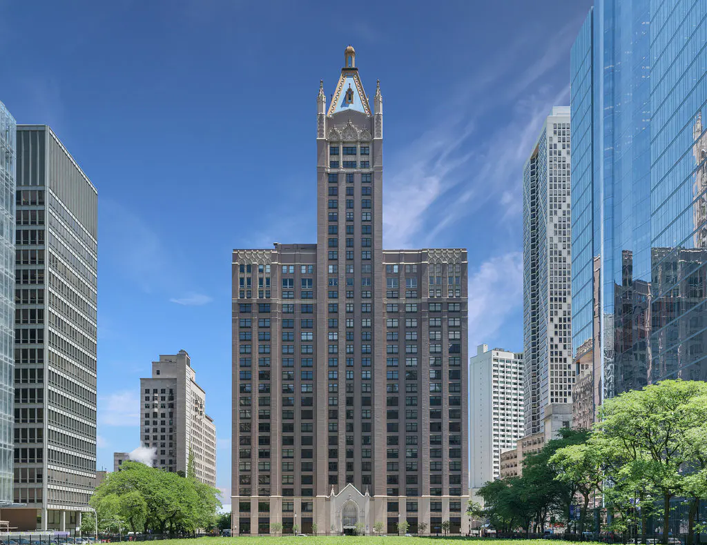 Exterior view of a tall, brown brick building with a pointed, ornate top, surrounded by modern glass skyscrapers and green trees under a blue sky.