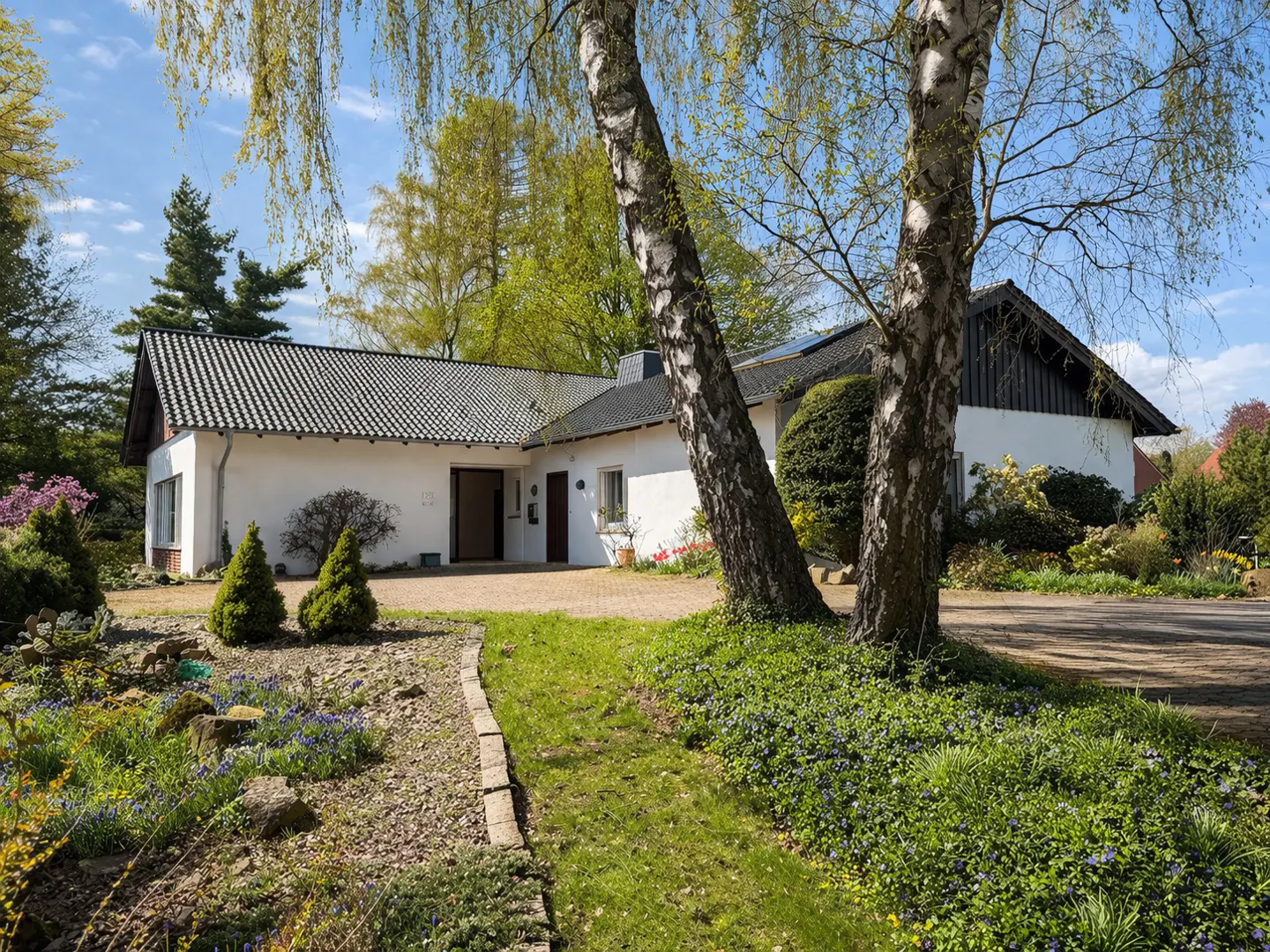 Exterior view of a white house with a dark gray roof, surrounded by trees and a garden with blue flowers.