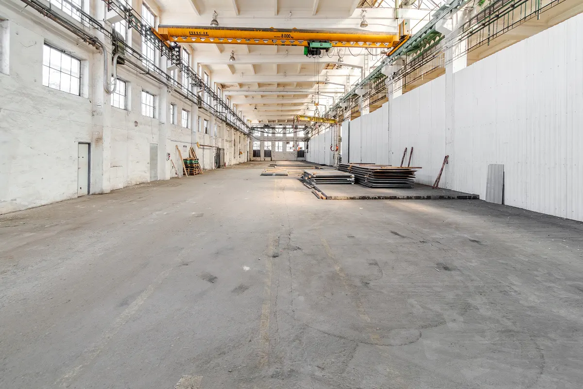 A large, empty warehouse with a concrete floor, white walls, and an overhead crane. Windows line the left wall.