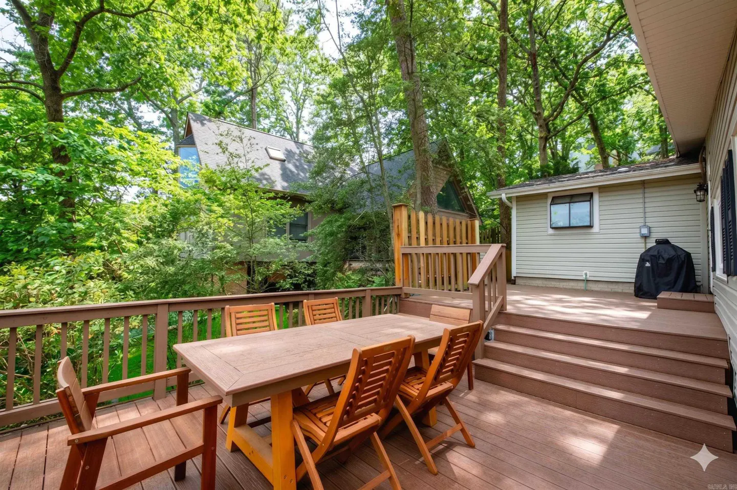 Wooden deck with table and chairs, surrounded by trees. A house is visible in the background. Grill on the side.