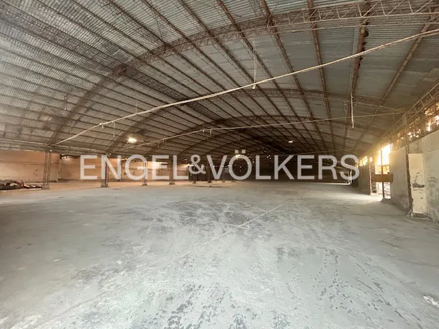 An empty warehouse with a gray concrete floor and a corrugated metal roof. The Engel & Völkers logo is superimposed on the image.
