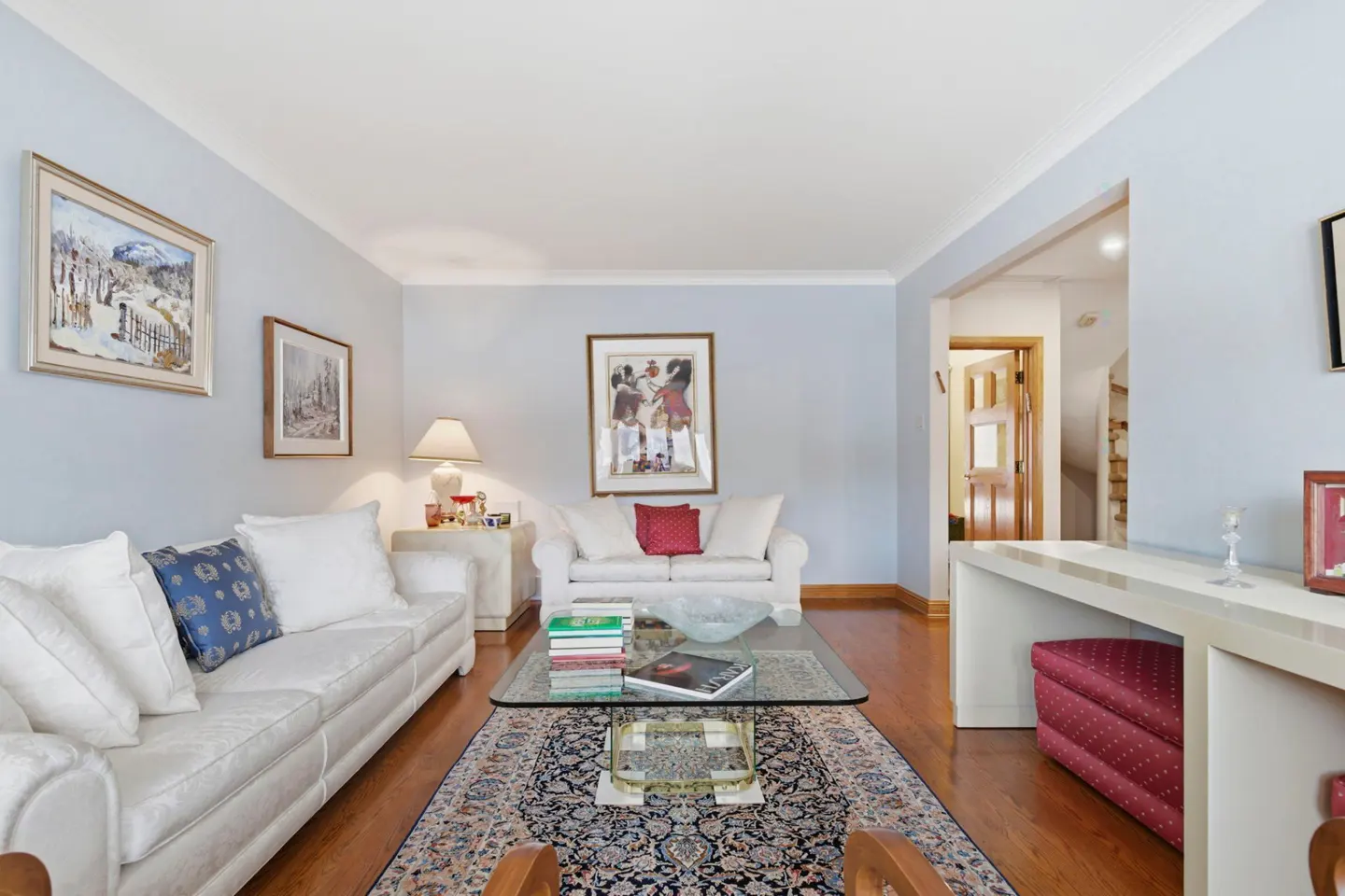 Living room with light blue walls, hardwood floors, and a patterned rug. Two white sofas face a glass coffee table with books. Artwork hangs on the walls.