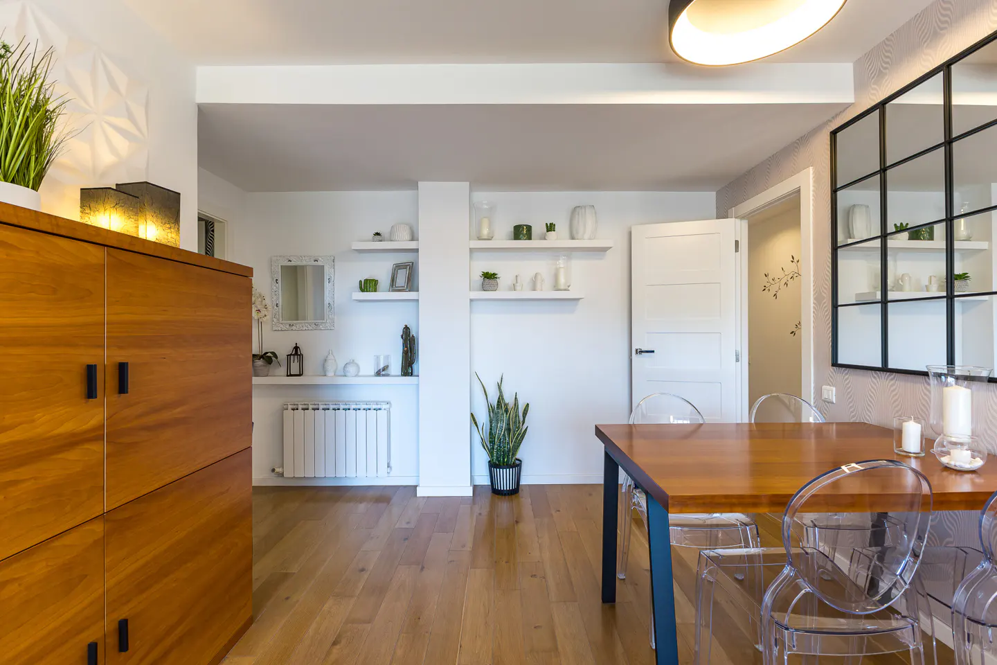 Bright dining room with wood table, clear chairs, and wood floor. White walls have shelves with decor. A wood cabinet stands to the left.