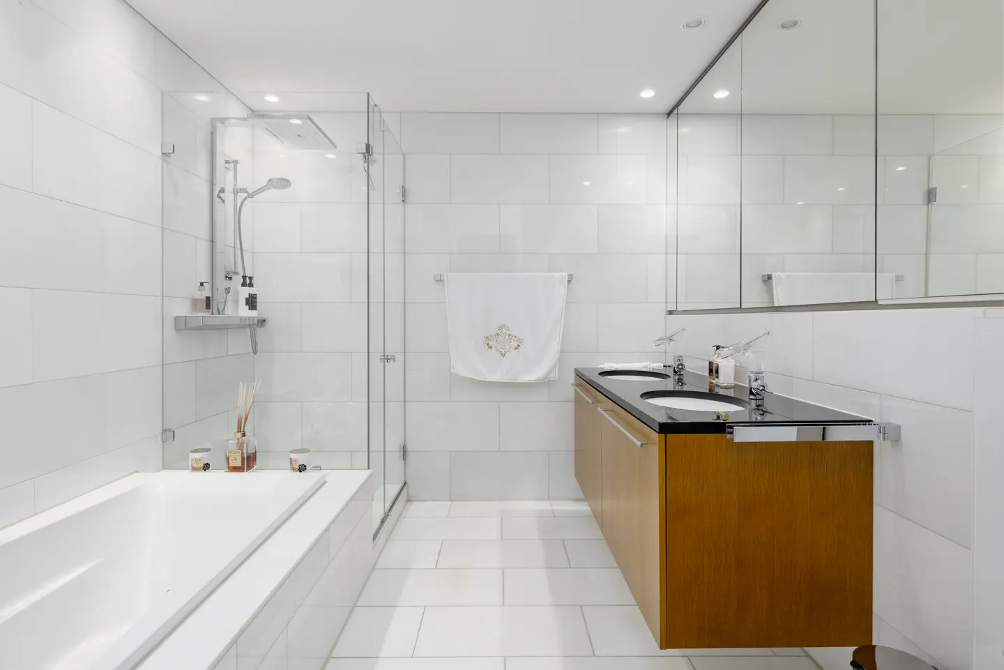 Modern bathroom with white tiled walls, a glass-enclosed shower, a white bathtub, and a floating wood vanity with black top.