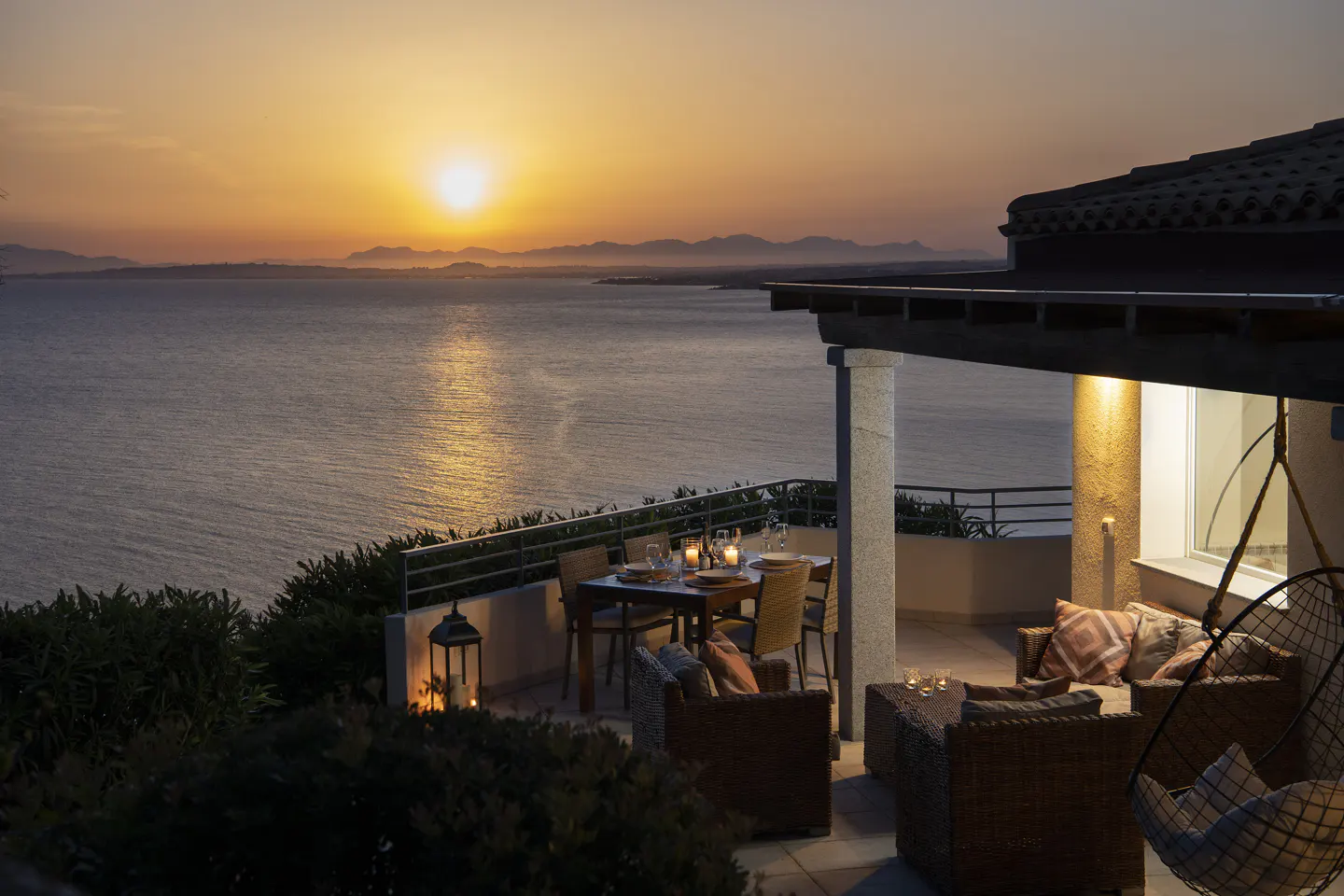 Sunset view from a villa patio with a dining table, wicker furniture, and a hanging chair overlooking the ocean.