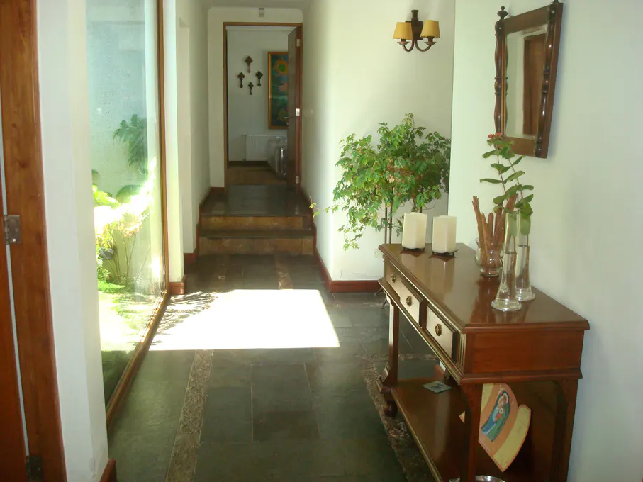 Hallway with dark stone floors, white walls, and a wooden console table with candles and vases. A glass wall shows a garden.