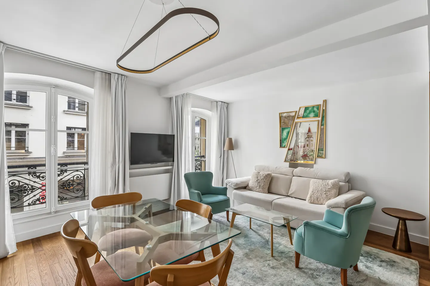 Bright living room with glass table, wood chairs, beige sofa, and teal armchairs. Windows show a Parisian building exterior.