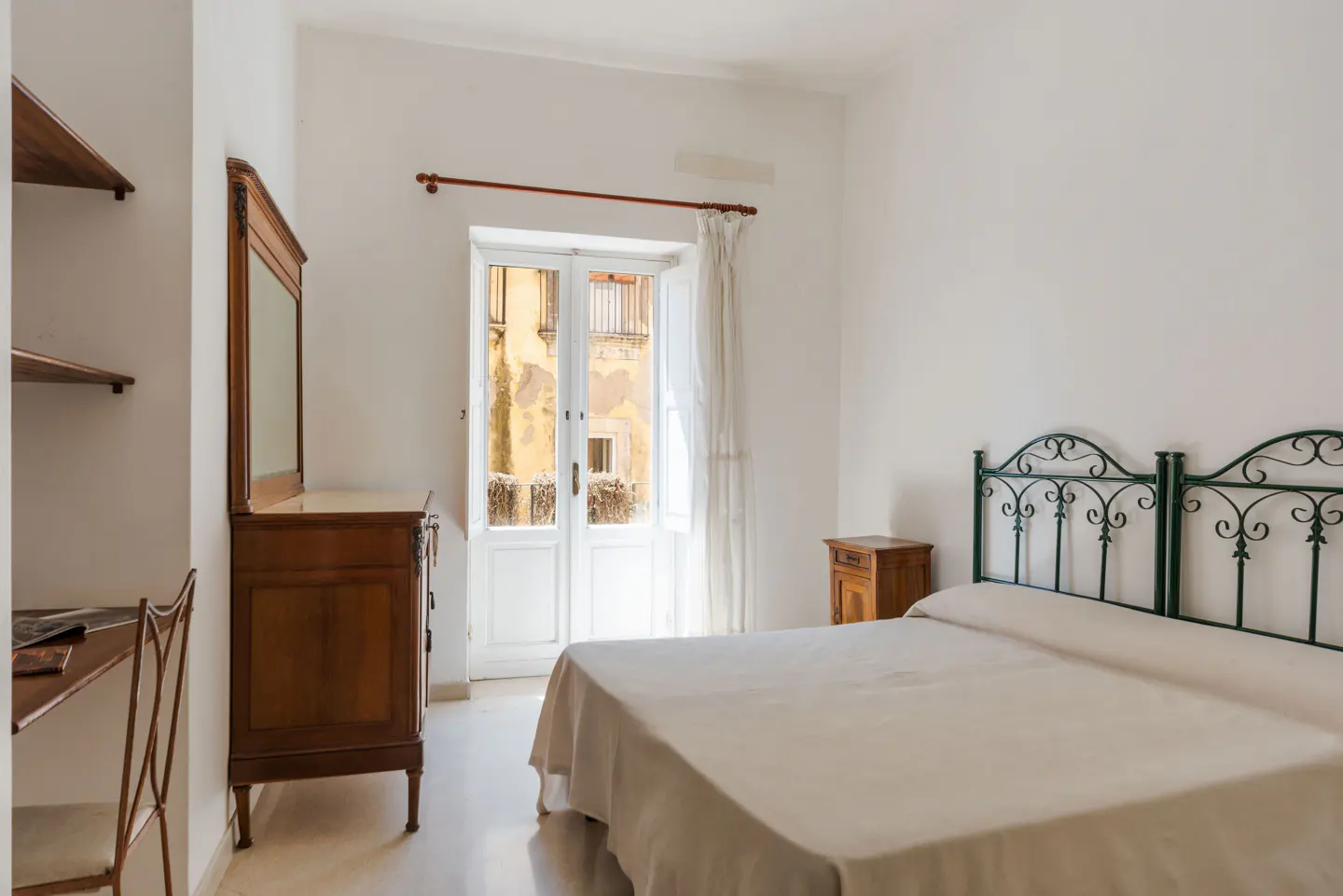 Bedroom with white walls, a wrought iron bed, and a wooden dresser with a mirror. Balcony doors open to a view of a building.
