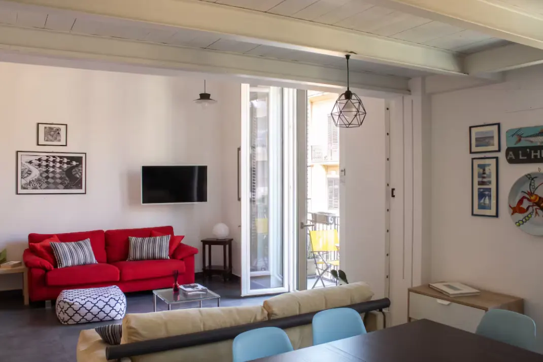 Bright living room with a red sofa, TV, and balcony with yellow chairs. White walls and ceiling beams.