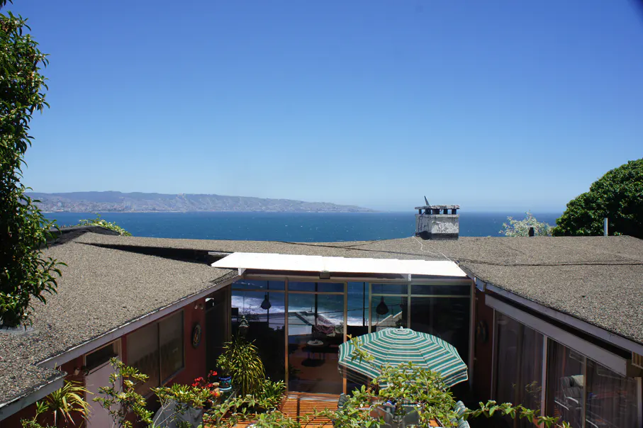 View of a house with a brown roof, a green and white umbrella, and a view of the ocean and mountains.