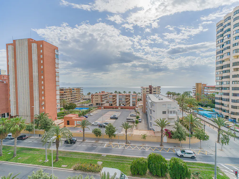 View of a coastal city with tall buildings, palm trees, and a parking lot. The ocean is visible in the background under a cloudy sky.