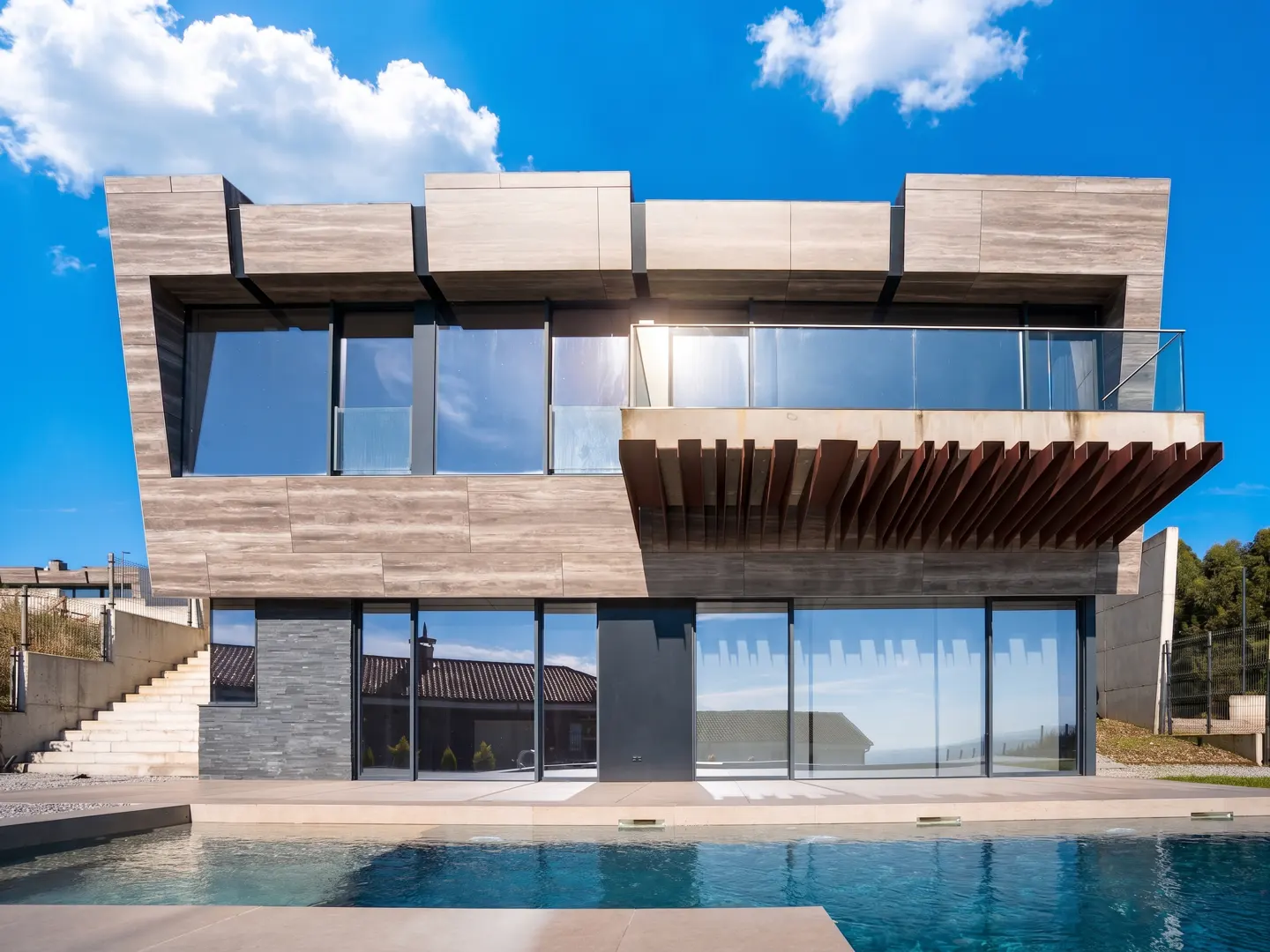 Modern two-story house with a pool. The house is gray with large windows and a wooden balcony. Blue sky with clouds.